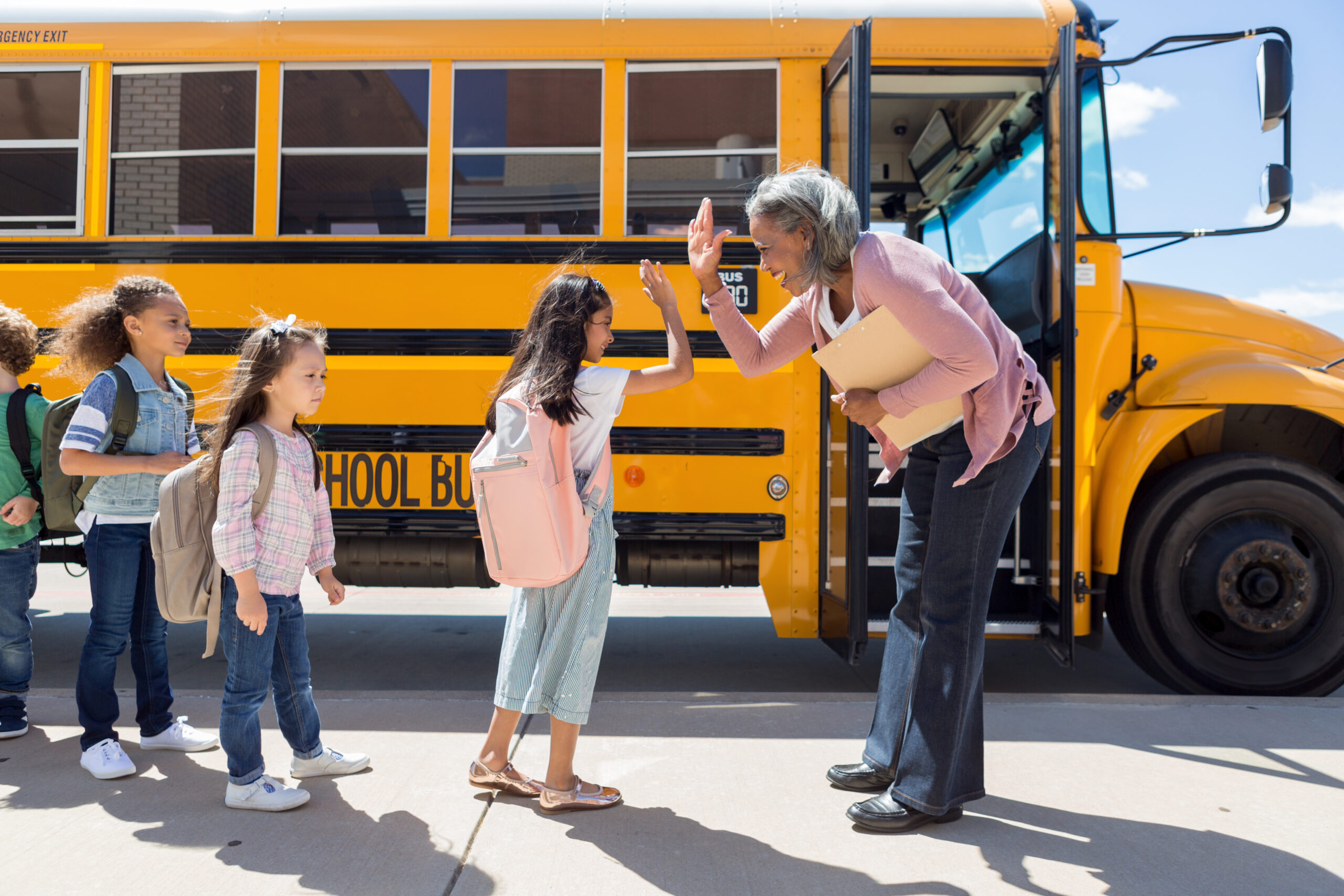 While waiting in line to board the school bus, a young female student gives the bus driver a high-five.