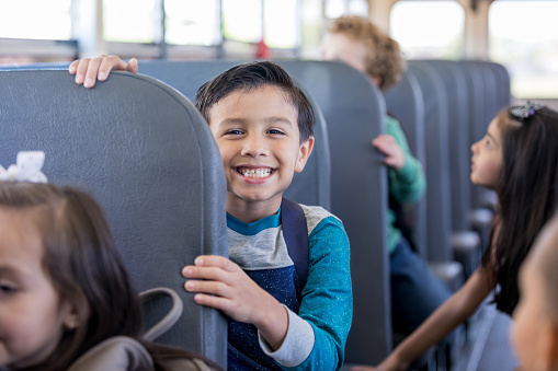 While sitting on the school bus with other children, a young schoolboy smiles with anticipation at the camera.